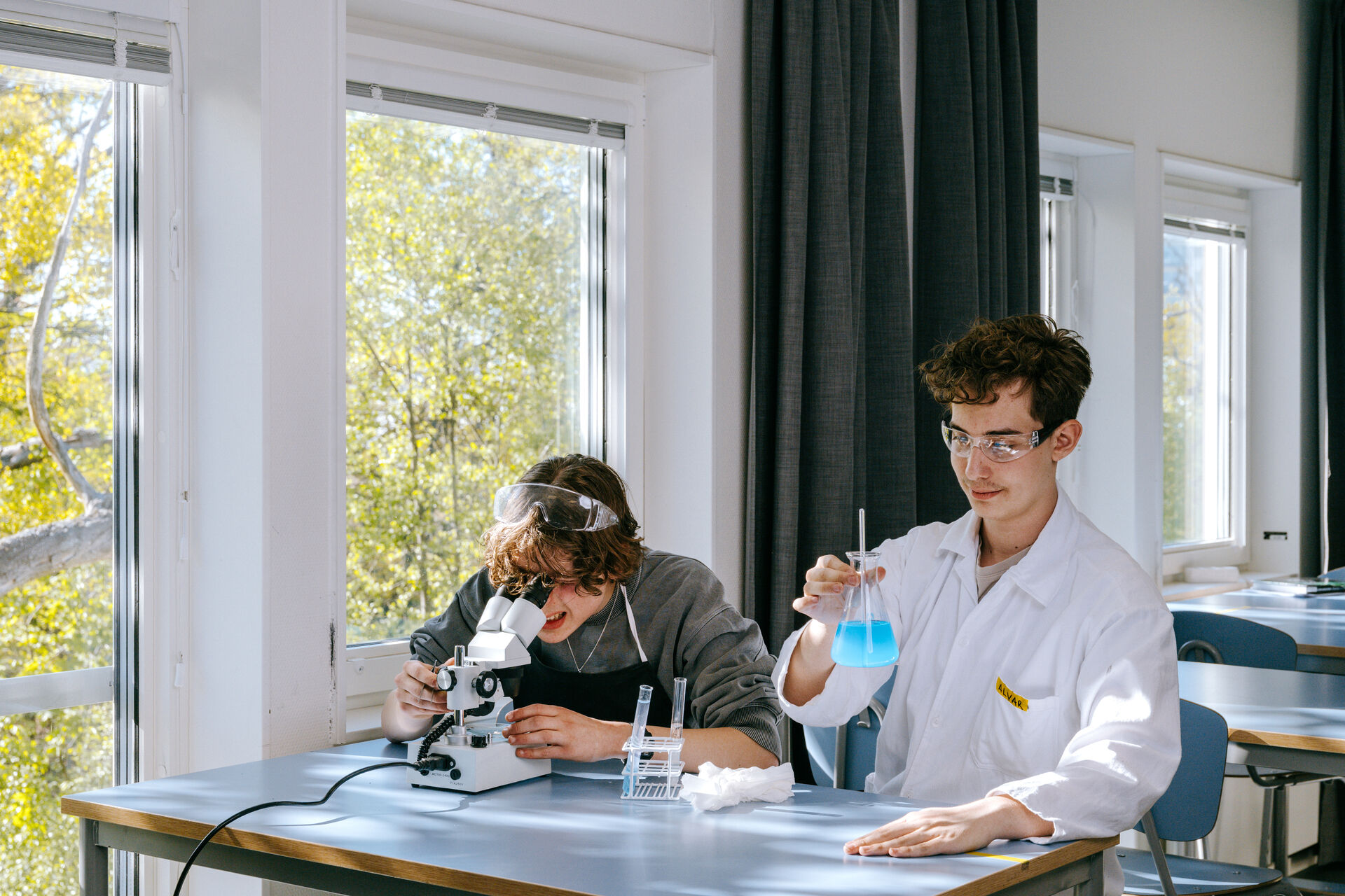 Två elever arbetar med naturvetenskapliga experiment i ett klassrum i Expandias modullösning på Ekens skola i Hammarby Sjöstad, där den ena eleven använder mikroskop och den andra blandar en blå vätska i laboratorieutrustning i en ljus och modern lärmiljö.