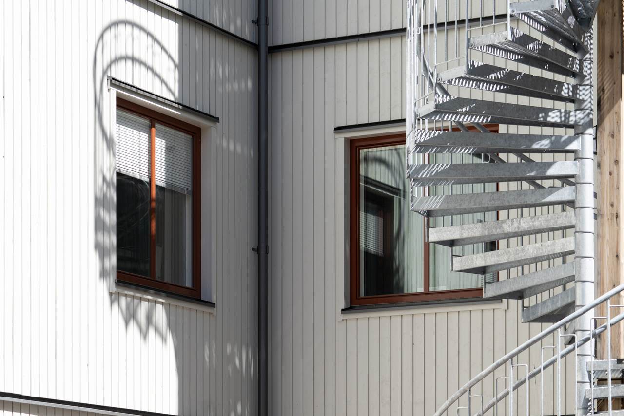 Detaljbild av kontorsmoduler från Expandia vid Centralsjukhuset i Karlstad. Fasaden i ljus kulör med vertikal panel och träinfattade fönster syns i närbild. En extern spiraltrappa i galvaniserat stål löper längs byggnaden och skapar tydliga skuggspel mot fasaden, vilket visar modulernas funktionella och genomtänkta arkitektur anpassad för sjukhusmiljö.