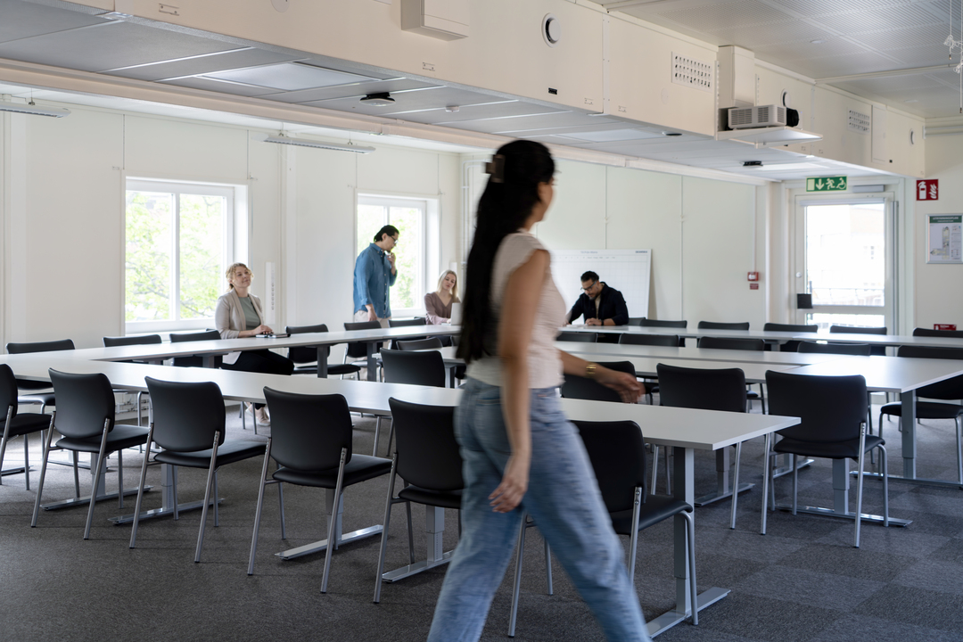 Stort konferensrum i kontorsmoduler från Expandia i Skanskas projektkontor i Karlstad. Långa bord och stolar är uppställda för möten, flera personer sitter samlade längre in i rummet och en person går förbi i förgrunden. Rummet är ljust med fönster som släpper in dagsljus.