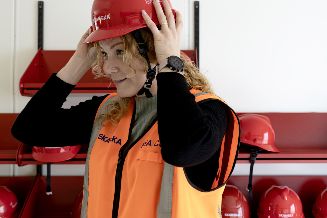 Person i Skanskas projektkontor i Karlstad som tar på sig en röd skyddshjälm. Personen bär varselväst och står framför ett ställ med skyddshjälmar i ett utrymme i kontorsmoduler från Expandia, anpassat för besökare och säkerhetsutrustning.