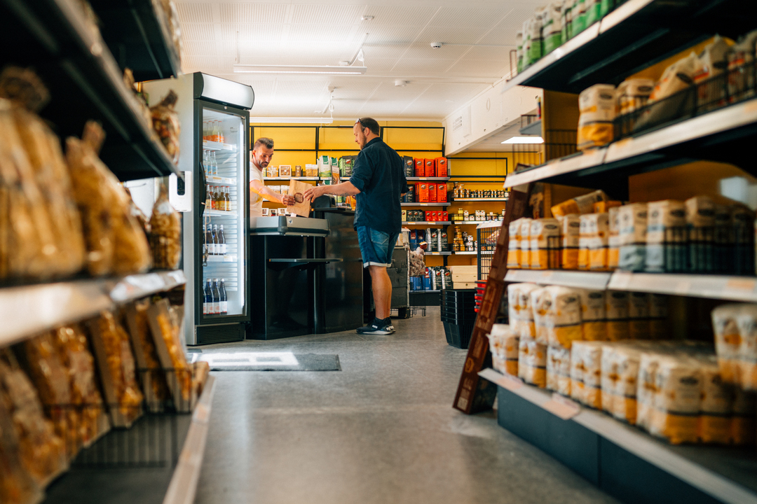 En man står vid kassan i en liten livsmedelsbutik och betalar, medan en butikspersonal tar emot varor över disken. Bilden är tagen från en låg vinkel mellan två hyllrader fyllda med förpackade livsmedel, och i bakgrunden syns kyldiskar och välfyllda hyllor i en ljus, modern butiksmiljö. Mannen befinner sig på Slemmestad centrum i Norge.
