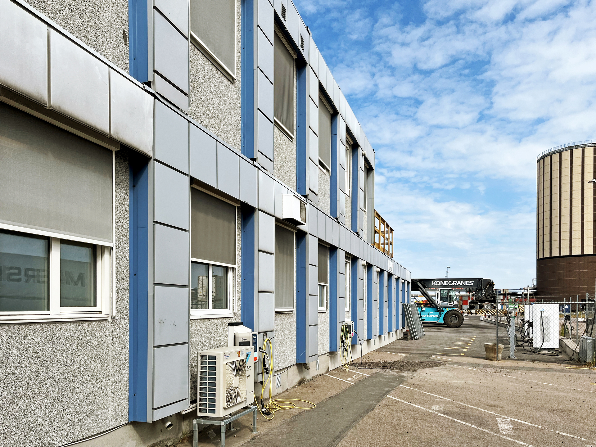 Lång rad av sammanbyggda moduler i Helsingsborgs hamn i grått i ett industriellt hamnområde, med fönster, externa ventilationsenheter och en asfalterad yta framför; i bakgrunden syns en hög cylindrisk industribyggnad under delvis molnig himmel.