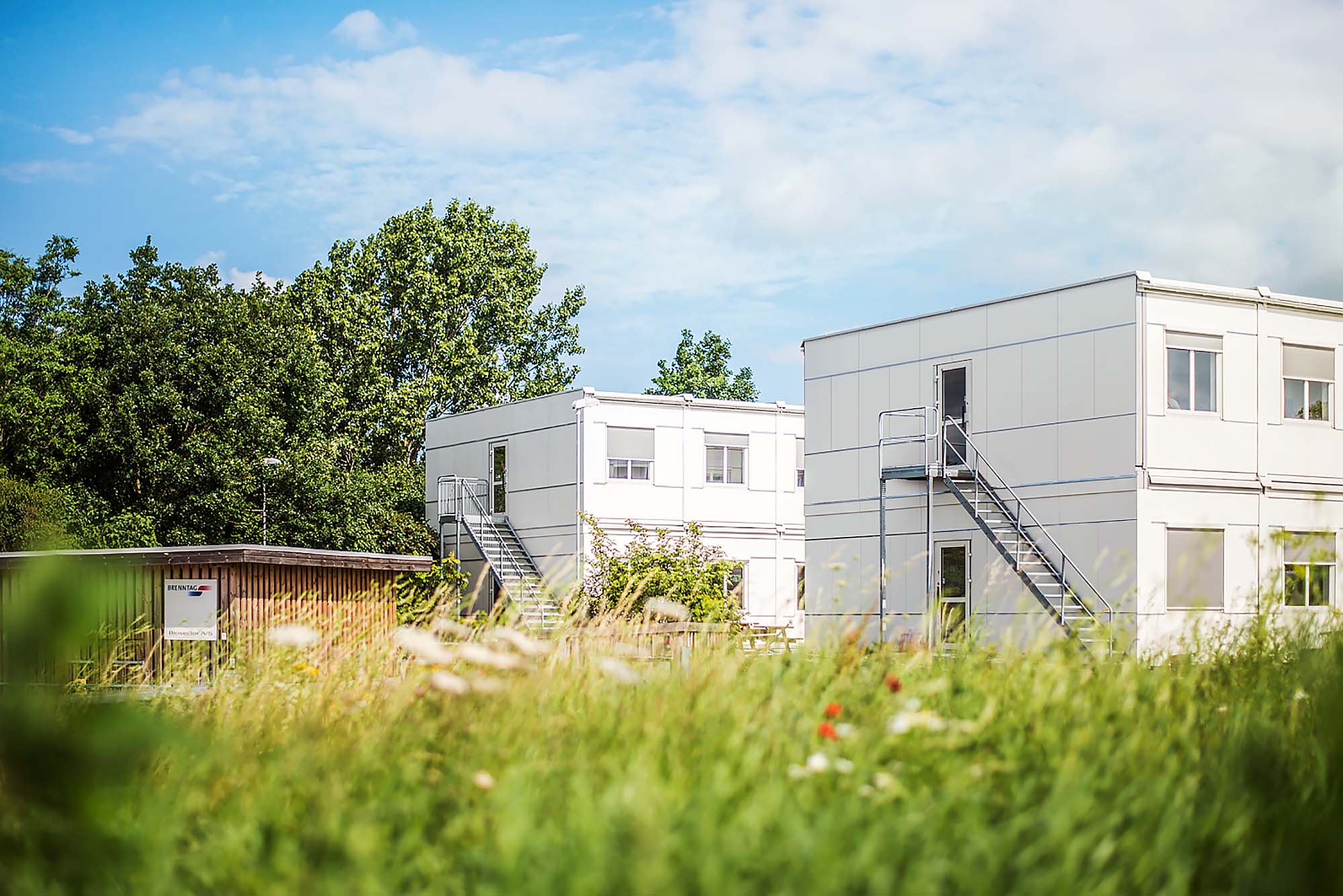 Vita kontorsmoduler i två plan placerade i grön utemiljö med träd och gräsytor. Flexibla kontorsmoduler från Expandia.