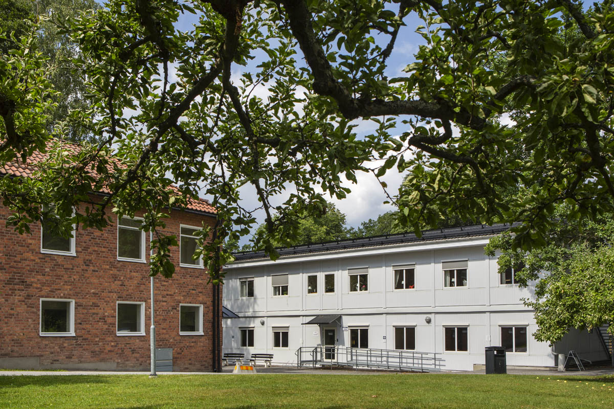 Ljusa kontorsmoduler i två plan med fönsterrader, entré med ramp och räcke, omgivna av grönska och träd i en lugn utemiljö – kontorsmoduler från Expandia.