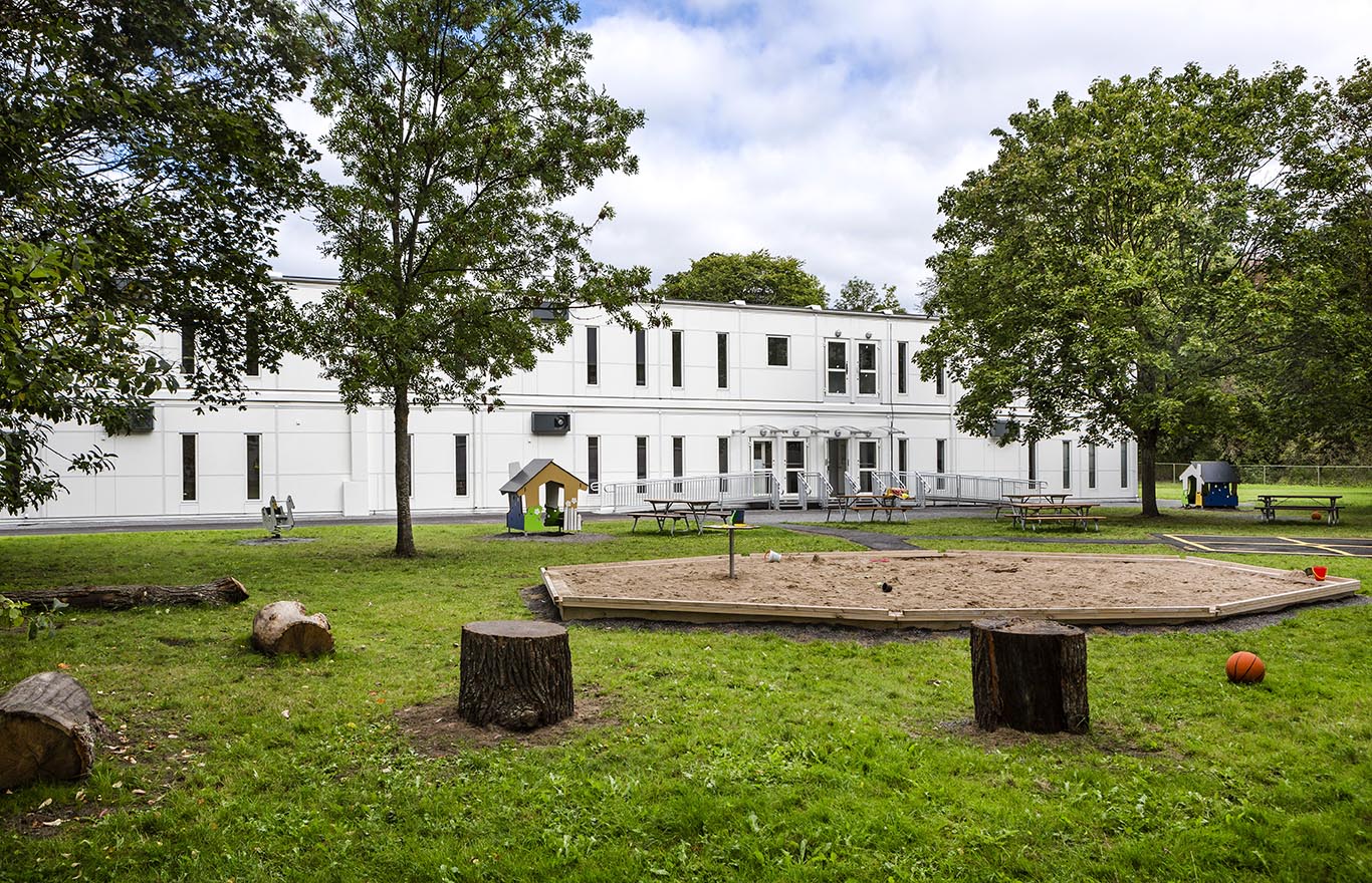 Exteriör vy av Sveaplansskolan i Stockholm, uppförd med skolmoduler från Expandia. Den vita tvåvåningsanläggningen omges av grönska med träd, gräsmatta och en skolgård med sandlåda, lekutrustning och sittplatser. I förgrunden syns en tillgänglighetsanpassad entré med ramp. Miljön upplevs som öppen, trygg och väl anpassad för skolbarn.