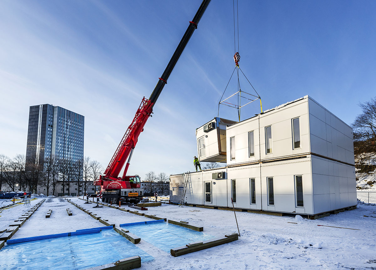 Montering av skolmoduler från Expandia vid Sveaplansskolan i Stockholm. En mobilkran lyfter en vit modul på plats ovanpå redan installerade moduler i ett vinterlandskap. På marken syns förberedda fundament och skyddsmaterial, medan en montör arbetar uppe på modulerna. I bakgrunden syns stadsmiljö med höghus och träd under klar blå himmel.