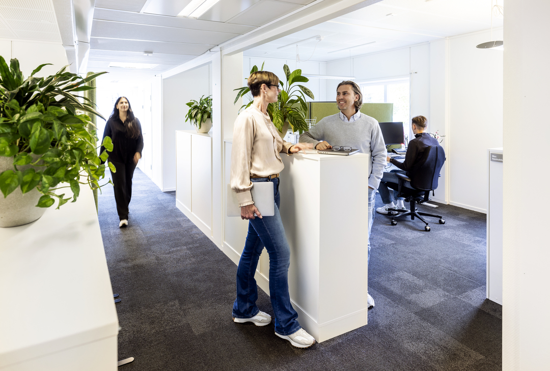 Ljus kontorsmiljö i kontorsmoduler hos Polypeptide där medarbetare samtalar vid en ståyta, med öppna arbetsplatser, gröna växter och mattbelagda golv som skapar en modern och trivsam arbetsmiljö – kontorsmoduler från Expandia.