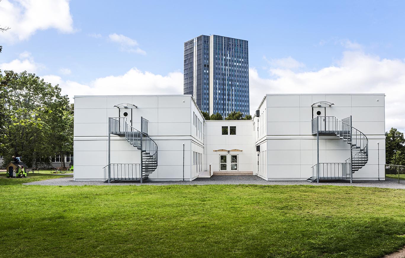 Skolmoduler från Expandia vid Sveaplansskolan i Stockholm. Två vita modulvolymer i två plan står placerade på en grön skolgård med utvändiga spiraltrappor. Mellan modulerna finns en entré, och i bakgrunden syns ett höghus samt omgivande träd under en ljus och molnig himmel.