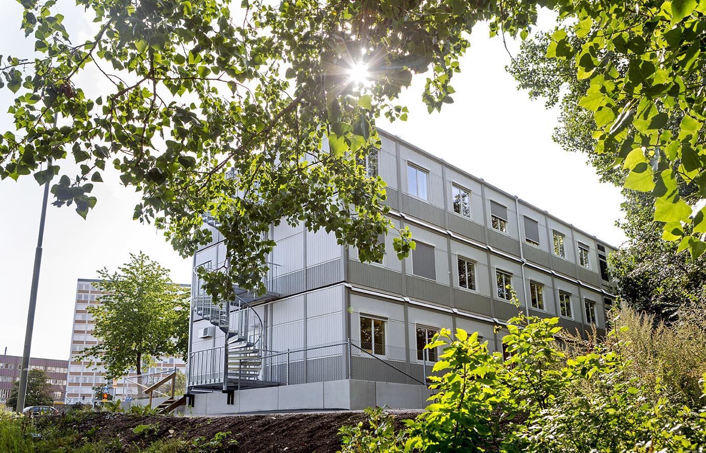 Tillfällig modulbyggnad från Expandia i tre våningar med kontorslokaler, placerad i grön stadsmiljö, med ljus fasad, många fönster och externa trappor.