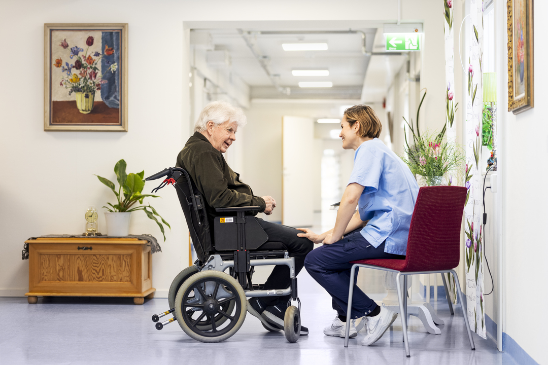 En boende i rullstol samtalar med omsorgspersonal i en korridor vid Borggården demensvård i Olofström. Den ljusa inomhusmiljön ingår i en modullösning från Expandia.