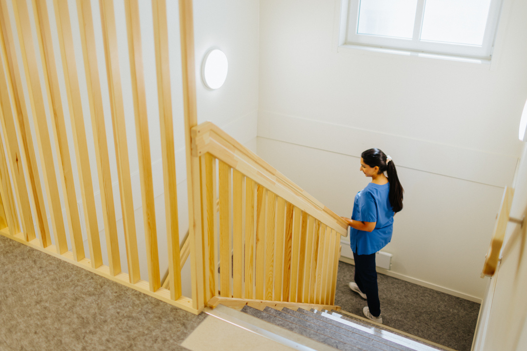 Omsorgspersonal går i en trappa med trähandledare inomhus vid Herrhagsgården vård- och omsorgsboende i Falun. Trapphuset har ljusa ytor och fönster som släpper in dagsljus.