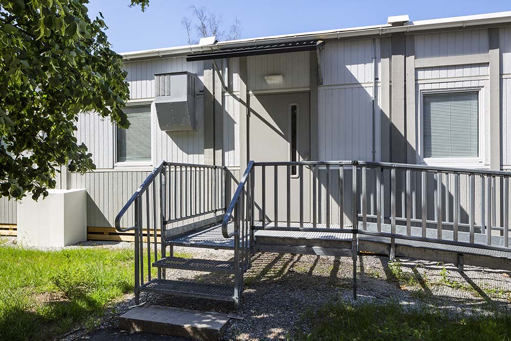 Exteriör av evakueringsboende i modulbyggnad med ljus fasad, entré med ramp och trappräcke, dörr med skärmtak samt fönster. Grönska och grusad mark runt byggnaden.