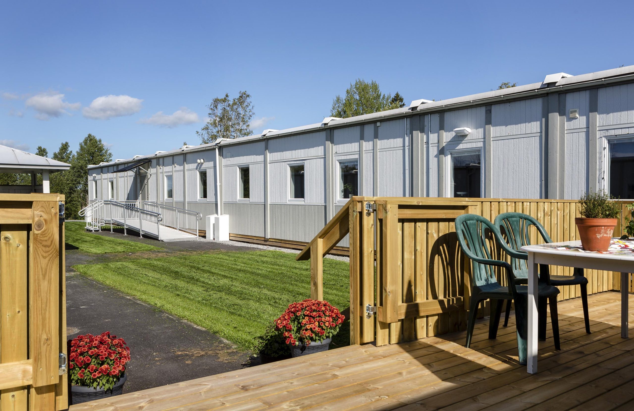 Uteplats vid Nordrona LSS-boende i Norrtälje med trädäck, bord och stolar, blomkrukor samt boendemoduler från Expandia i bakgrunden och tillgänglig ramp längs fasaden.