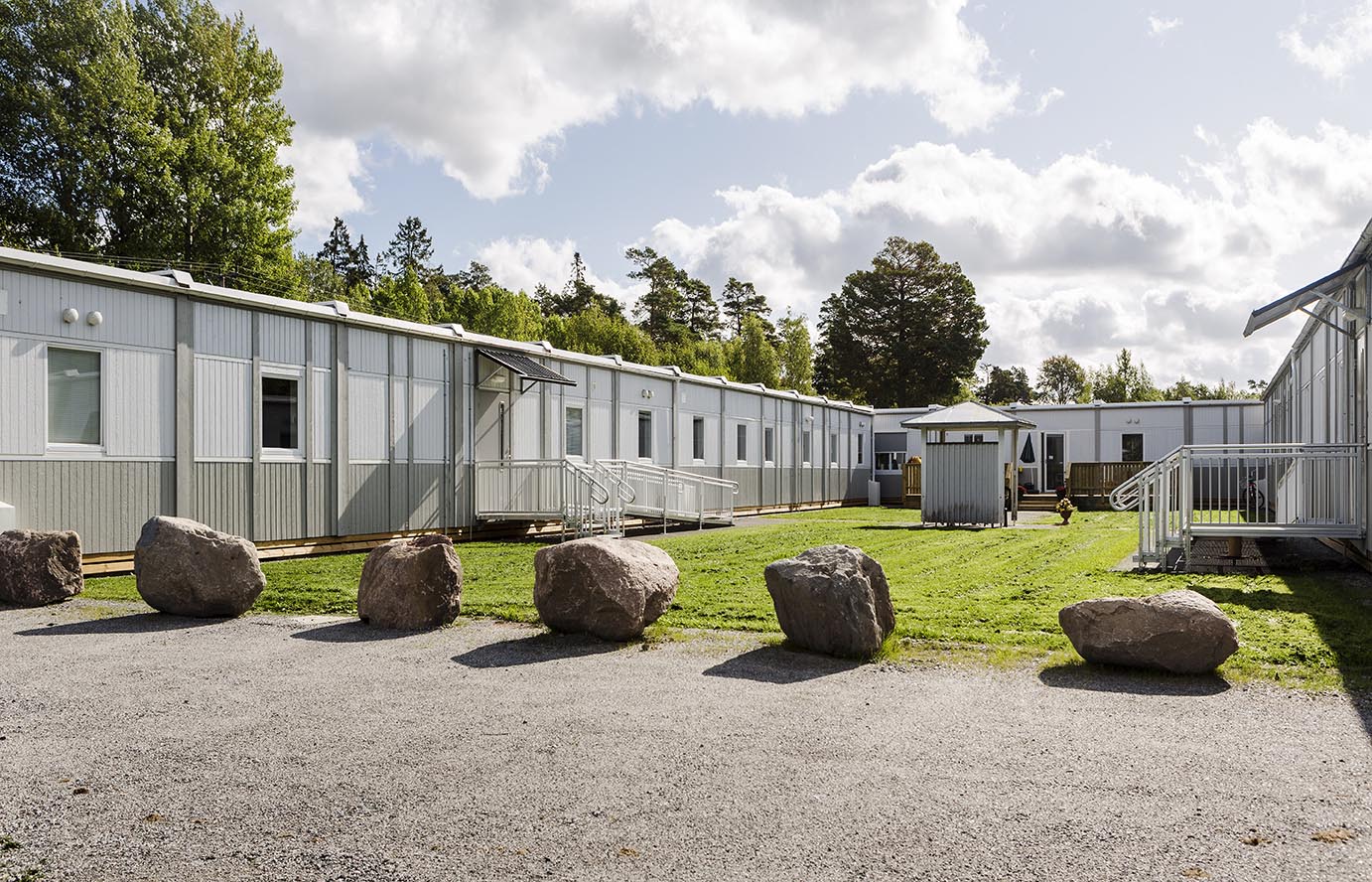 Nordrona LSS-boende i Norrtälje med ljusa boendemoduler från Expandia placerade runt en grön innergård, med ramper, grusgångar och stora stenar i förgrunden.